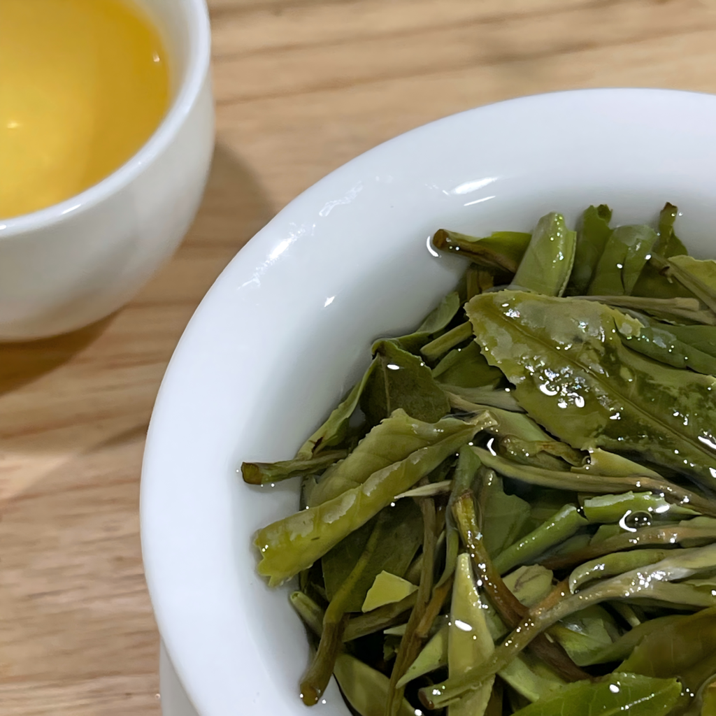A close-up view of premium Bai Mu Dan white tea, highlighting the brilliant, pale golden-amber liquor in a clear glass cup and the unfurled spring leaves.